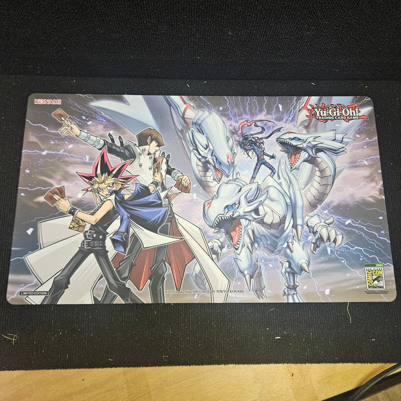 A Yu-Gi-Oh! trading card playmat featuring artwork of the White Eyed dragon and Yugi and Seto Kaiba, displayed on a black mousepad over a wooden surface.